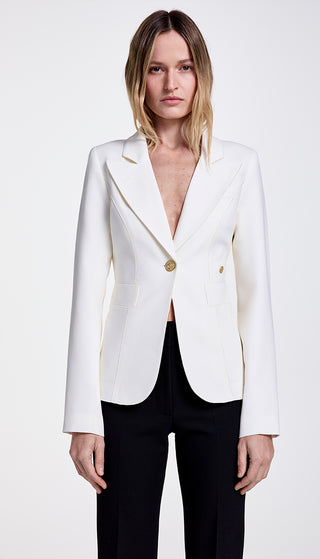 A model wearing a white single-breasted blazer with a cut-out back vent and brass hardware, paired with black pants.