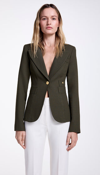 A woman wearing a green single-breasted blazer with a cut-out back vent and brass hardware, paired with white trousers.