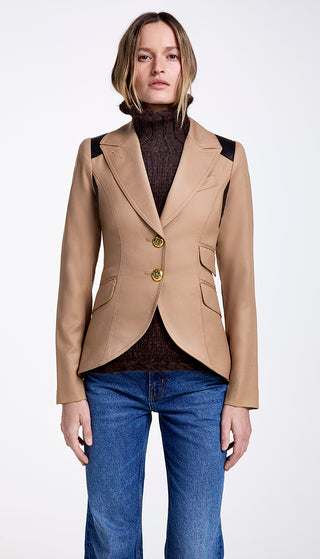 A woman modeling a beige camel twill equestrian blazer with black leather detailing and brass buttons, paired with a dark top and blue jeans.