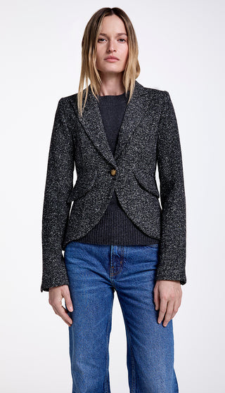 A woman wearing a black and gray tweed one button blazer with rectangle elbow patches, paired with a black top and blue jeans.