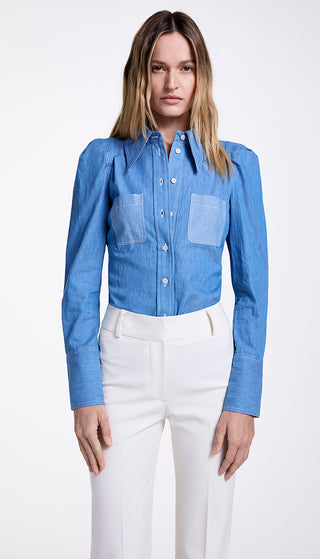 A woman wearing a blue denim shirt with a point collar and box pleat shoulders, featuring tonal patch pockets, paired with white trousers.
