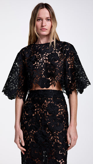 A model wearing a black boxy top with guipure lace detailing, paired with a matching skirt. The top features a dropped armhole, voluminous sleeve, and lace-edged hem.