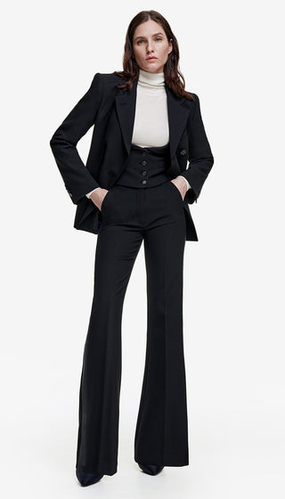 A model wearing a high waisted tuxedo pant with a wide leg and a pocket on the side, featuring a tonal tuxedo stripe. The outfit is black piqué.