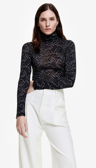 A person wearing a black turtleneck with a geometric pattern, paired with white trousers.