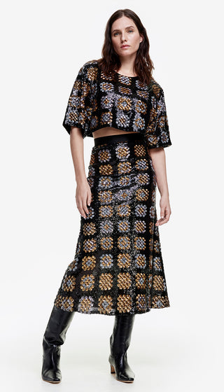 A model wearing a black and gold sequin midi skirt with a geometric pattern, paired with a matching top and chunky knit sweater.