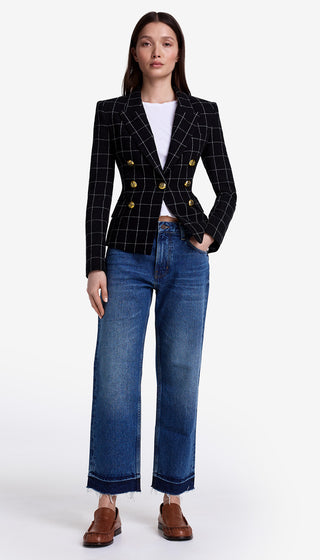 Woman wearing a black checkered blazer with gold buttons, white shirt, blue jeans, and brown shoes on a white background.