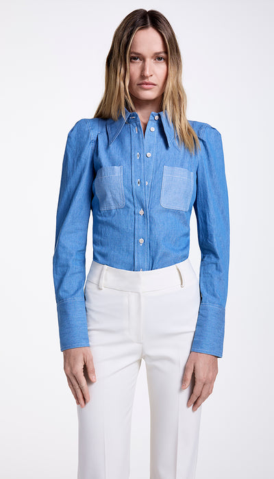 A woman wearing a blue denim shirt with a point collar and box pleat shoulders, featuring tonal patch pockets, paired with white trousers.