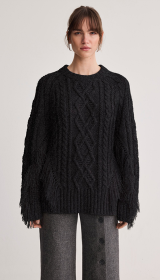A woman modeling a black cable-knit sweater with fringe detailing on the sleeves, paired with gray button-front trousers, against a plain light gray studio background.