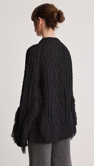 A woman shown from the back wearing a black cable-knit cardigan with fringe trim, paired with gray pants, against a plain light gray studio background.
