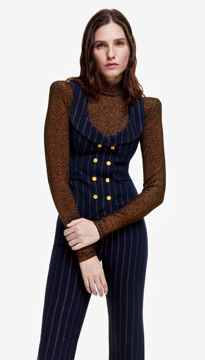 A woman wearing a double-breasted, navy pinstripe waistcoat with a portrait neck, paired with a brown long-sleeved top and trousers.