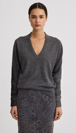 A woman modeling a dark gray long-sleeve V-neck knit sweater with a matching gray lace skirt against a light gray studio background.