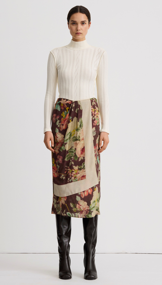 A model stands against a plain light gray studio background wearing a cream ribbed mock-neck long-sleeve top, a wrap-style midi skirt with a floral print and beige border, and black knee-high boots.