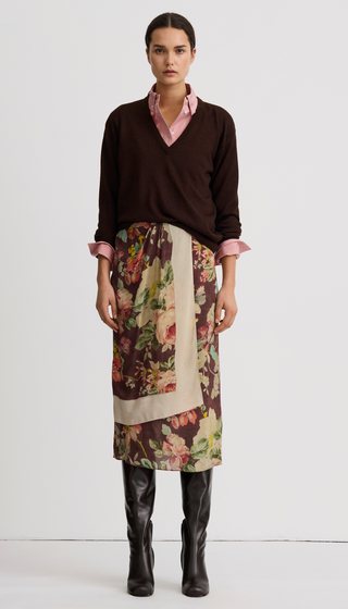 A model stands against a plain light gray studio background wearing a dark brown V-neck sweater over a pink collared shirt, a floral wrap-style skirt with beige panels, and black knee-high boots.