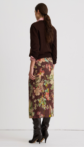 A model shown from the back wearing a dark brown long-sleeve knit top, a brown midi skirt with a colorful floral print, and black knee-high heeled boots, against a plain white studio background.