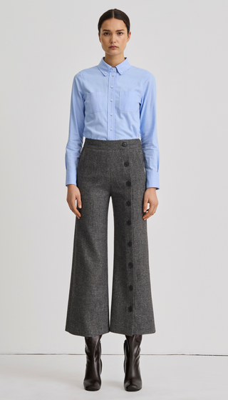 A model stands against a light gray studio background wearing a light blue long-sleeve button-up shirt tucked into high-waisted dark gray wide-leg trousers with a front button placket, paired with black ankle boots.