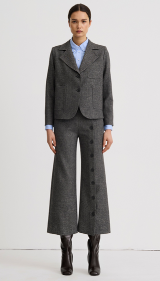 A model stands against a plain light gray studio background wearing a tailored gray suit with a cropped blazer, matching wide-leg trousers with front buttons, a light blue collared shirt, and black ankle boots.