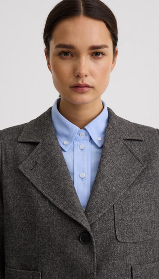 A close-up of a model wearing a textured gray wool-blend blazer over a light blue button-down shirt, set against a plain light gray background.