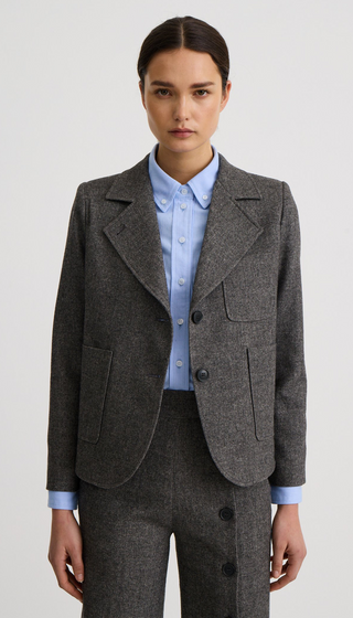 A model wearing a structured gray tweed blazer over a light blue button-down shirt, paired with matching gray trousers, against a plain light gray studio background.
