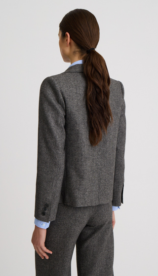 A woman shown from the back wearing a tailored gray tweed blazer and matching trousers over a light blue collared shirt, against a plain light gray studio background.