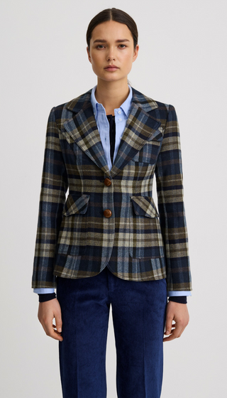 A model wearing a tailored plaid blazer in navy, beige, and brown tones, layered over a light blue button-up shirt and paired with navy trousers, shot against a plain light gray studio background.