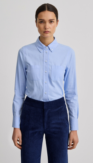 A woman modeling a light blue long-sleeve button-up shirt with two chest pockets, paired with dark navy corduroy trousers, against a plain light gray studio background.
