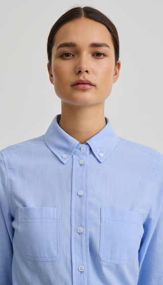 A woman wearing a light blue long-sleeve button-down shirt with two chest pockets, photographed against a plain light gray studio background.