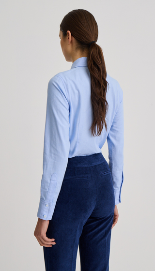 A woman shown from the back wearing a light blue long-sleeve button-up shirt and dark navy trousers, photographed against a plain light gray studio background.