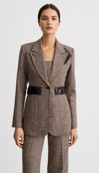 A model wearing a tailored brown tweed blazer and matching trousers with black satin lapels and a matching black waist belt, photographed against a light gray studio background.