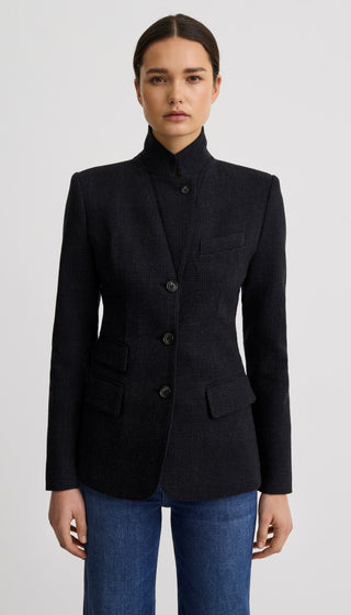 A woman modeling a dark navy blue textured blazer with a high stand collar and three front buttons, paired with blue jeans, against a light gray studio background.