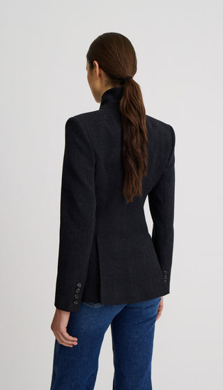 A woman viewed from the back wearing a dark charcoal gray structured blazer with a high mock neck and blue jeans, set against a plain light gray studio background.