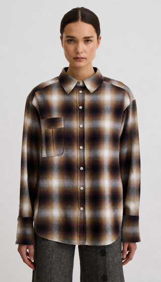 A model wearing a long-sleeve button-up plaid shirt in brown, beige, and black tones, paired with gray trousers, against a plain light gray background.