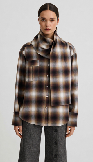 A model wearing a brown, beige, and black plaid button-up shirt with a matching oversized cowl neck scarf, paired with dark gray wide-leg trousers, against a light gray studio background.