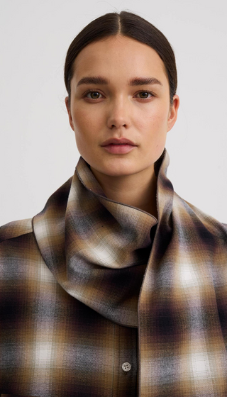A close-up portrait of a model wearing a brown, beige, and white plaid scarf wrapped around her neck, paired with a matching plaid button-up shirt against a light gray background.