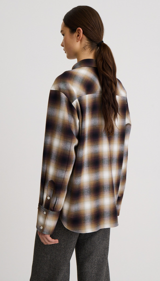 A woman shown from the back wearing an oversized long-sleeve plaid button-up shirt in brown, beige, and black tones, paired with dark gray pants, against a plain light gray studio background.