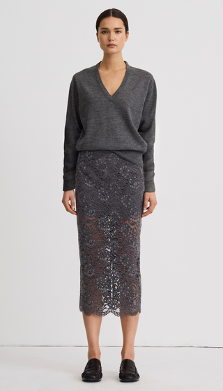 A model stands against a plain light gray studio background wearing a dark gray V-neck knit sweater and a matching dark gray lace midi skirt, paired with black loafers.