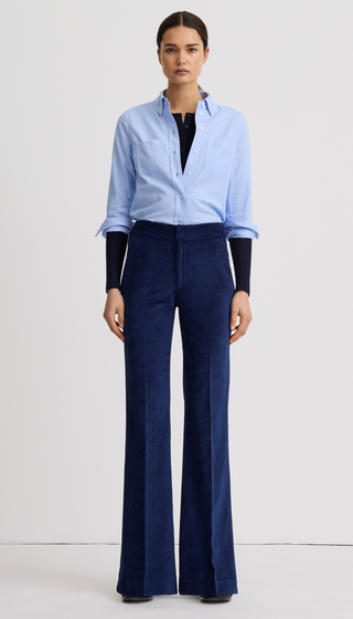 A model stands against a plain white studio background wearing a light blue button-up shirt with rolled sleeves, a black top underneath, and dark blue wide-leg trousers.