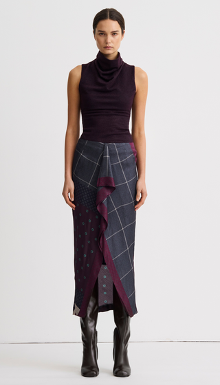 A model stands against a plain white studio background wearing a dark plum sleeveless cowl-neck top and a long wrap-style skirt with a dark gray and navy plaid pattern, deep purple ruffle accents, and small blue polka dots, finished with dark brown knee-high boots.