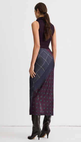 A woman shown from the back wearing a sleeveless dark plum top and a long patterned skirt with dark gray plaid and purple polka-dot panels, paired with dark brown knee-high boots, against a plain white studio background.