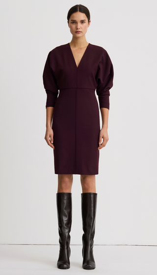 A model stands against a plain light gray studio background wearing a knee-length dark plum dress with a deep V-neck, long balloon sleeves, and black knee-high leather boots.