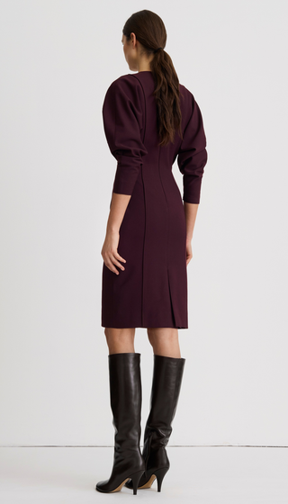 A woman shown from the back wearing a knee-length dark plum dress with puffed three-quarter sleeves and black knee-high heeled boots, photographed against a plain light gray studio background.