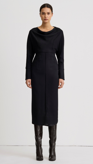 A model stands against a light gray studio background wearing a long-sleeve black cowl-neck midi dress with a defined waist and black knee-high boots.