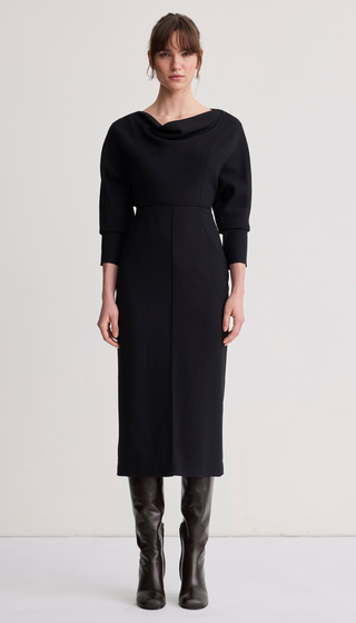 A model stands against a plain off-white studio background wearing a black cowl-neck midi dress with three-quarter sleeves and dark brown knee-high leather boots.