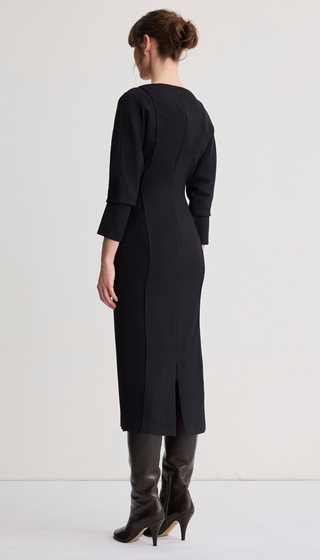 A woman shown from the back wearing a black knee-length dress with three-quarter sleeves and black heeled boots against a plain light studio background.