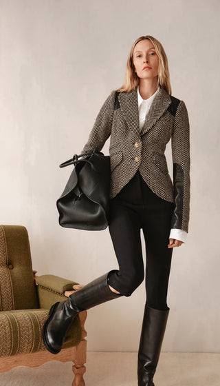 Woman in a patterned blazer and black pants holding a black bag in an indoor setting.