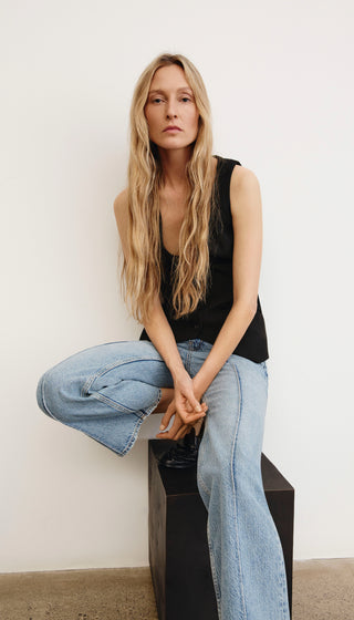 Woman sitting on a black stool wearing a black sleeveless top and light blue jeans against a white wall.