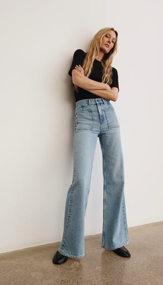 Woman wearing a black top and light blue jeans against a white wall.