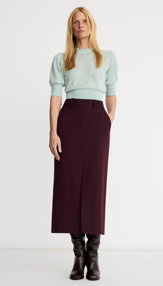 A woman stands against a plain white studio background wearing a pale mint green short-sleeve knit top with puffed shoulders and a dark burgundy midi skirt, paired with black ankle boots.