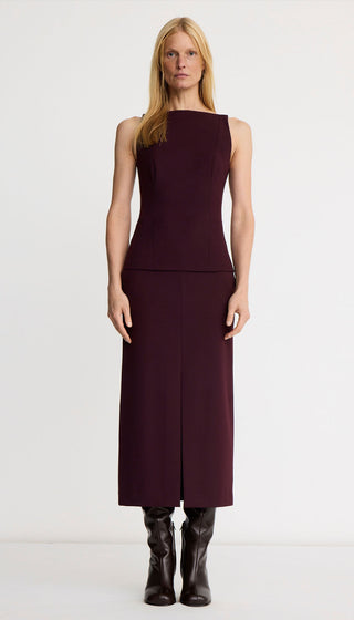A model stands against a plain white studio background wearing a sleeveless dark plum midi dress with a structured bodice and a front slit, paired with black ankle boots.