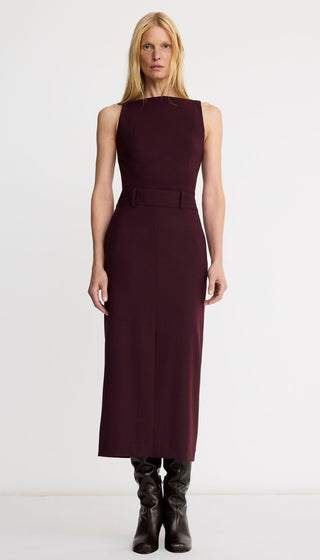 A blonde model wearing a sleeveless, high-neck, knee-length dark plum midi dress with a matching belt, paired with black leather boots, against a plain white studio background.