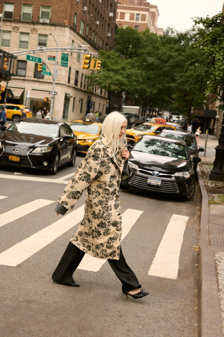 A woman with long platinum blonde hair walks across a city crosswalk wearing a long cream-colored coat with a dark floral pattern, black wide-leg pants, and black pointed shoes. The background shows a busy New York City street with cars, yellow cabs, and trees.